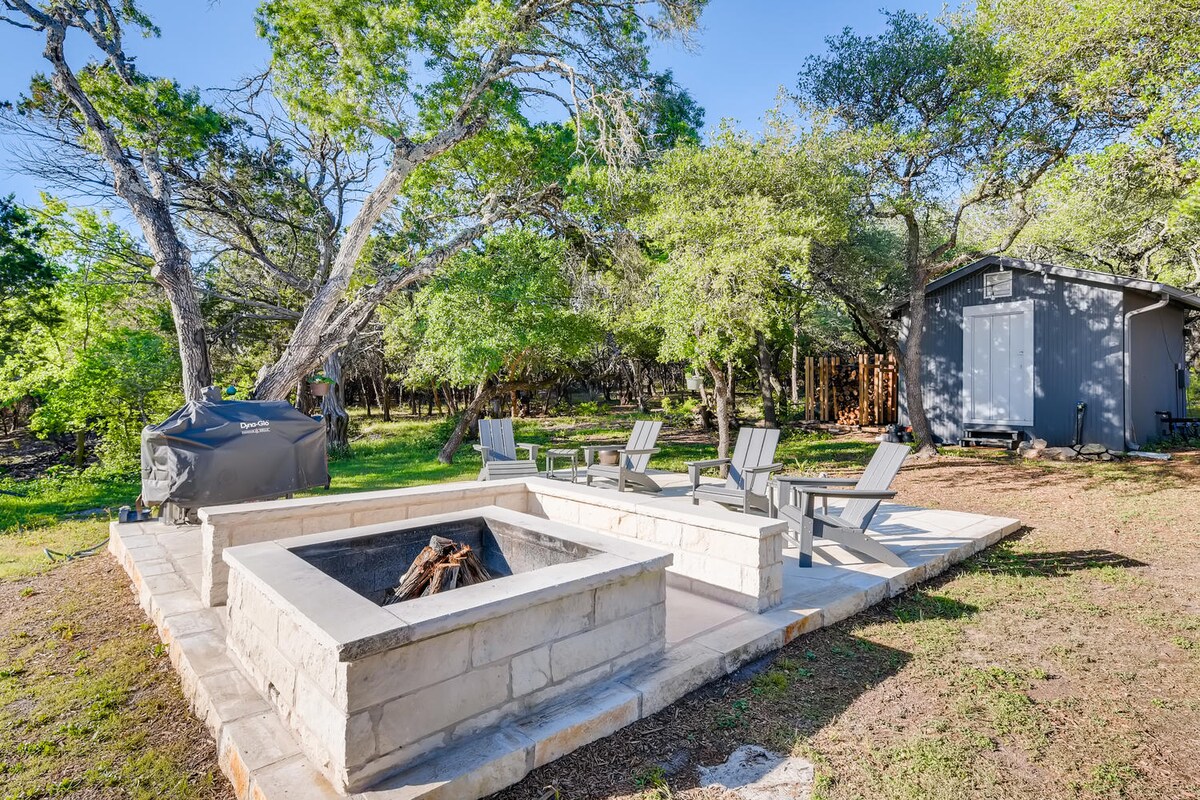Outdoor fire pit area near The Cabin at Eco Valley featuring a square stone fire pit, Adirondack chairs on a stone patio, grill cover, and a small outbuilding surrounded by trees in a peaceful woodland setting