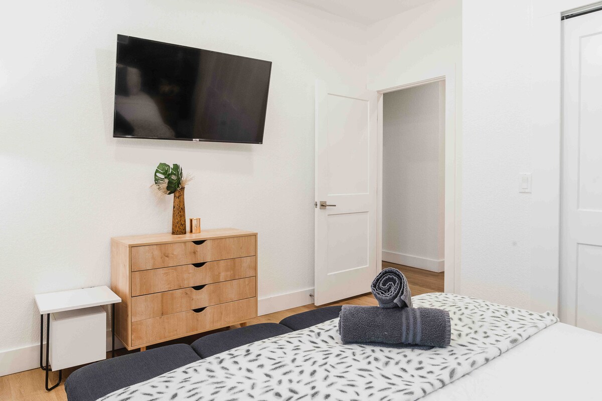 Bedroom at The Villa with wall-mounted TV, wood dresser, neatly styled bed with folded towels, minimalist decor, and bright neutral walls creating a calm, modern space