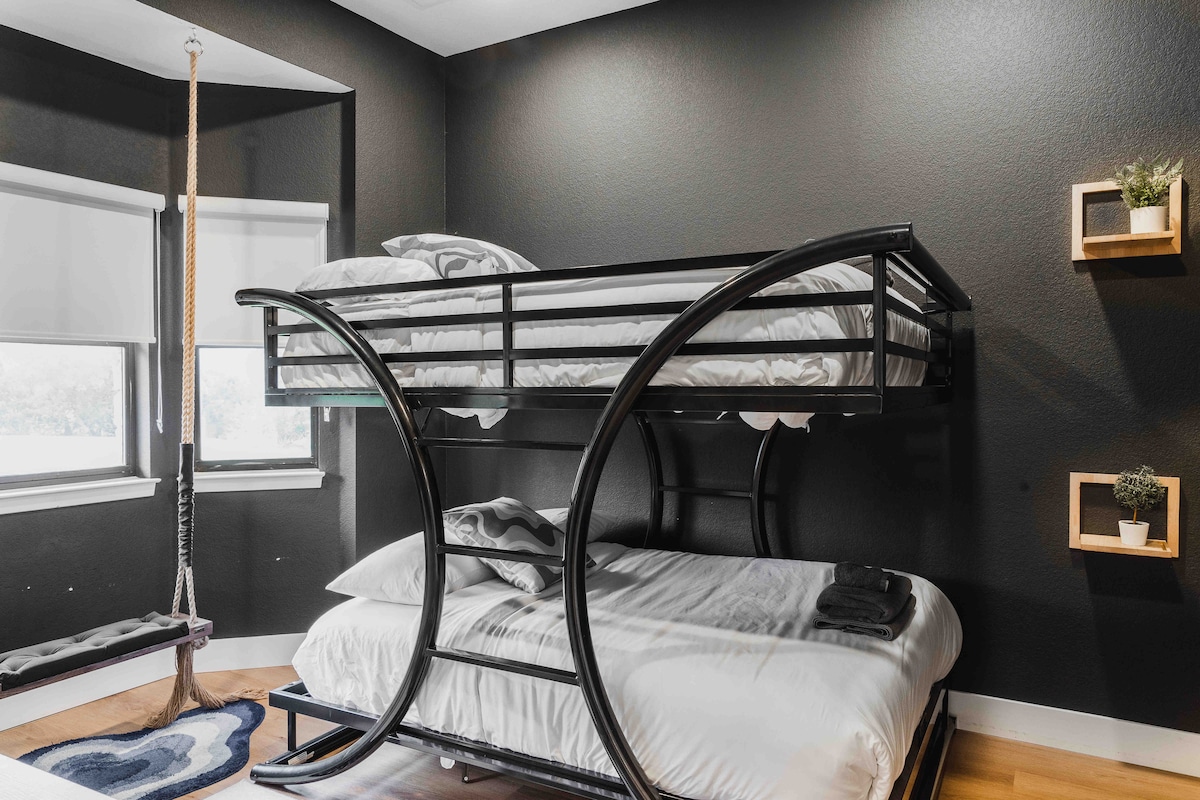 Bunk bedroom at The Villa with curved metal bunk beds, dark walls, white bedding, rope swing seat, small wall shelves, and warm wood flooring creating a modern, playful space