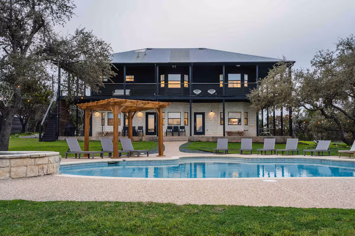 Front exterior view of The Villa showcasing a two-story stone and metal home, resort-style swimming pool, pergola seating, lounge chairs, and landscaped lawn framed by mature oak trees