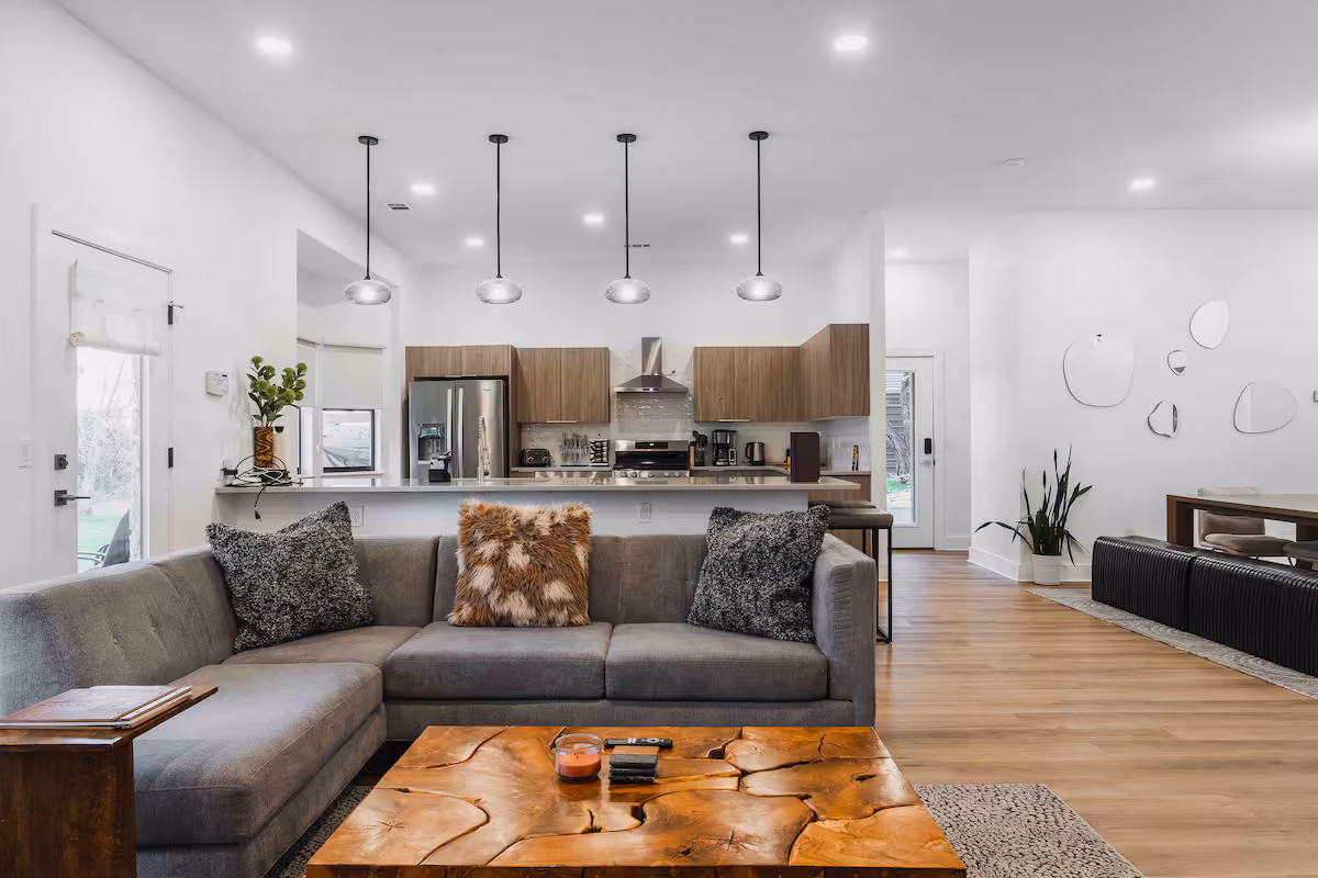 Open living area at The Villa featuring a modern sectional sofa, wood coffee table, pendant lighting, and a spacious kitchen with wood cabinetry and breakfast bar in a bright, airy layout