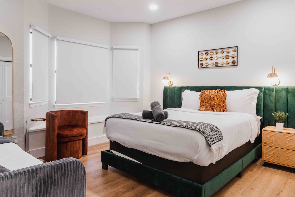 Bedroom at The Villa with green upholstered bed, white linens, accent pillows, bedside sconces, wood nightstand, cozy seating area, and large windows with soft natural light