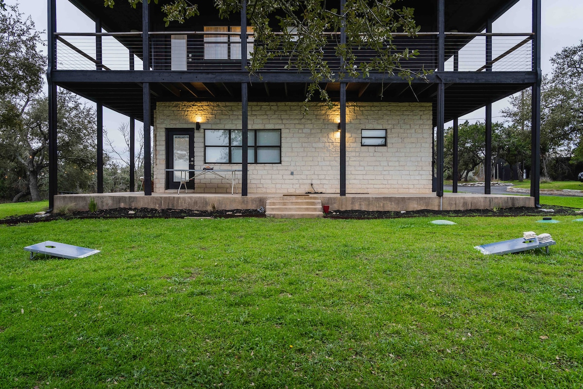 Rear exterior of The Villa featuring stone facade, covered patio with balcony above, green lawn, and outdoor cornhole boards beneath mature oak trees