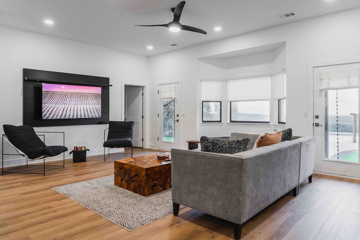 Modern living room at The Villa with gray sofa, wood coffee table, wall-mounted TV, lounge chairs, ceiling fan, and large windows overlooking hill country views