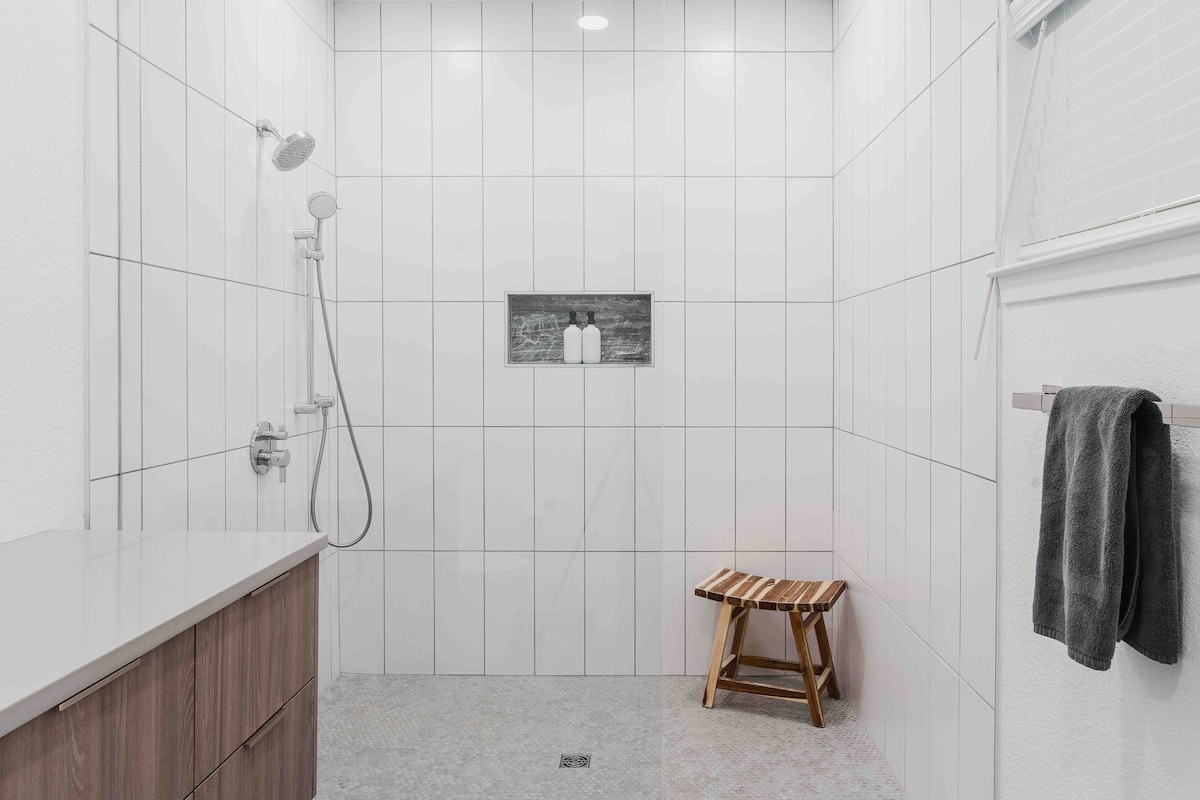 Walk-in shower in The Villa with white tiled walls, rainfall shower head, built-in niche, wooden stool, and modern minimalist design
