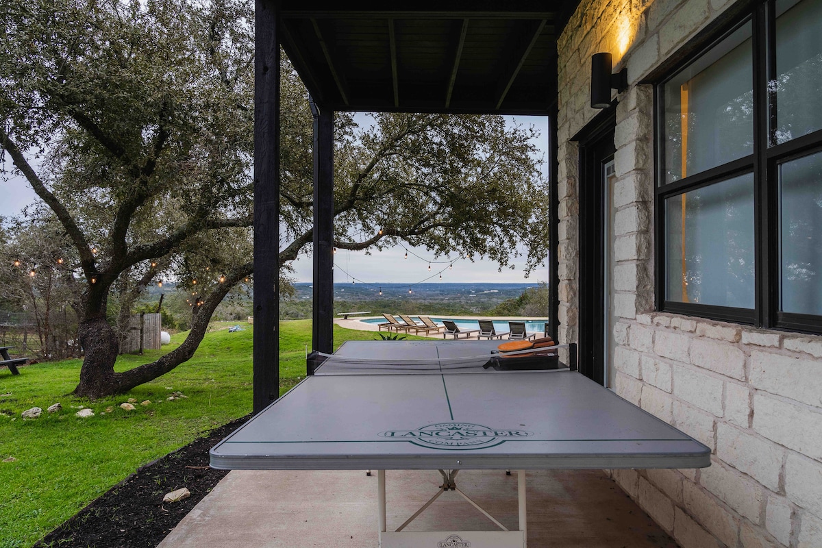 Covered patio at The Villa with an outdoor ping pong table, string lights, stone exterior, and scenic hill country views overlooking the pool