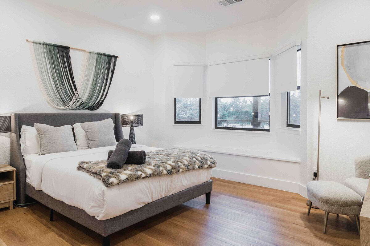 The Villa bedroom with a king bed, layered neutral bedding, soft accent chair, large windows with natural light, and warm wood flooring