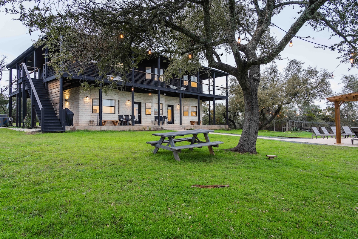 Outdoor lawn at the villa featuring a picnic table beneath mature oak trees, string lights overhead, and a modern two-story villa with wraparound balcony in the background.