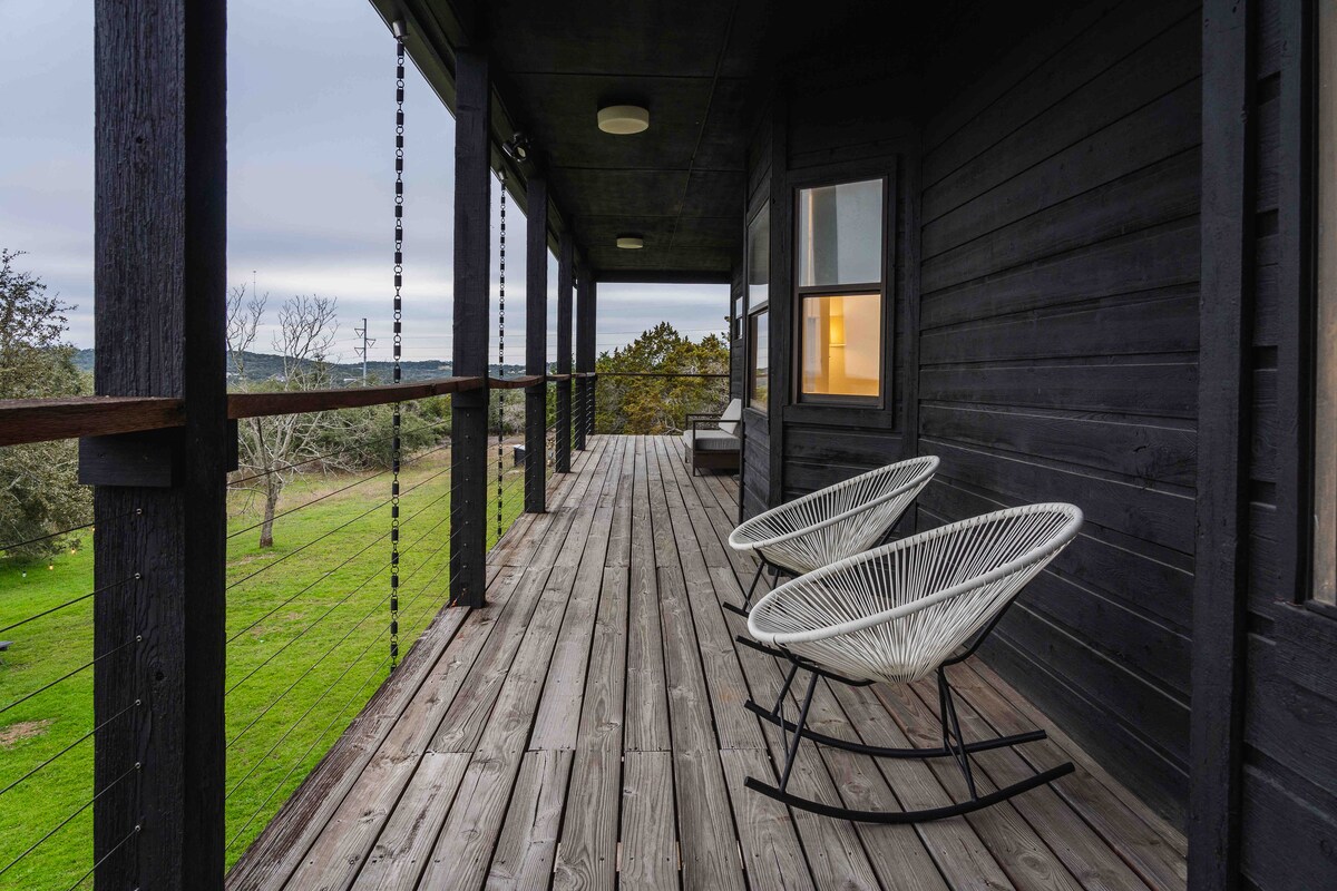 Covered upper balcony at The Villa with wood plank flooring, modern rocking chairs, cable railing, dark exterior siding, and peaceful hill country views overlooking green landscape