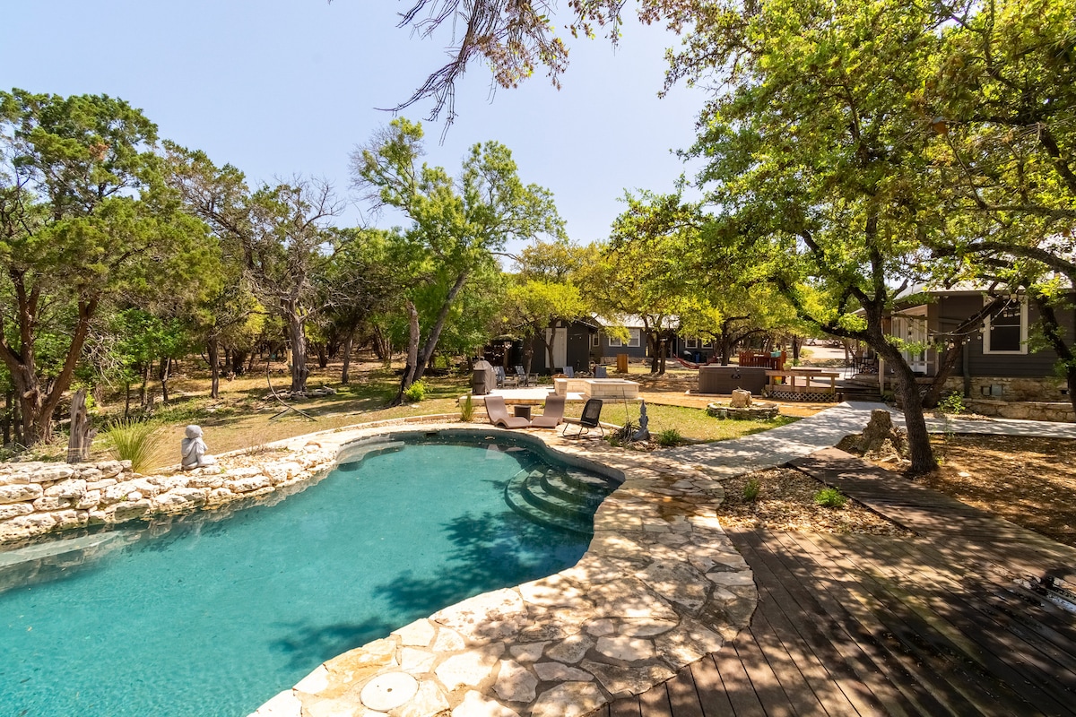 Wide view of The Nook at Eco Valley showing a natural stone swimming pool with curved steps, surrounding lounge chairs, fire pit area, and cabins nestled among mature trees in a serene woodland setting
