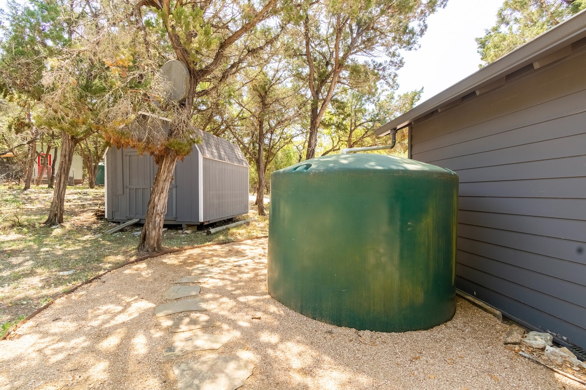 Rainwater collection tank at The Nook at Eco Valley beside a cabin and storage shed, highlighting sustainable water conservation in a wooded setting