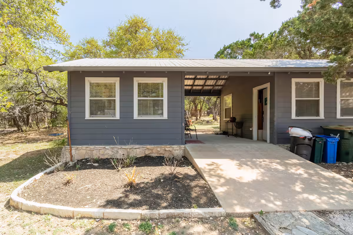 Front entrance of The Nook at Eco Valley showing a cozy cabin with covered porch, gray exterior, and surrounding trees in a peaceful woodland setting