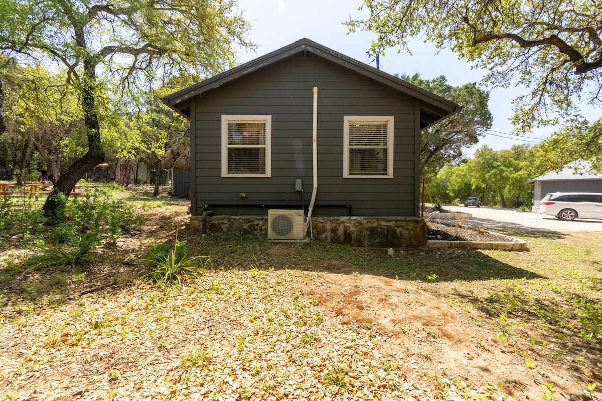 Exterior view of The Nook cabin at Eco Valley surrounded by trees, featuring a dark green building with two windows and a peaceful natural setting