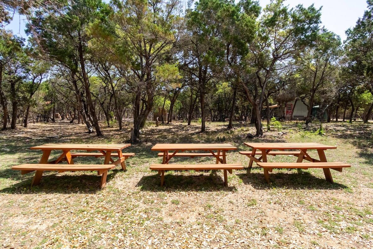 Multiple wooden picnic tables arranged in a forest clearing at the Nook, surrounded by cedar trees and natural landscape for outdoor gathering and dining