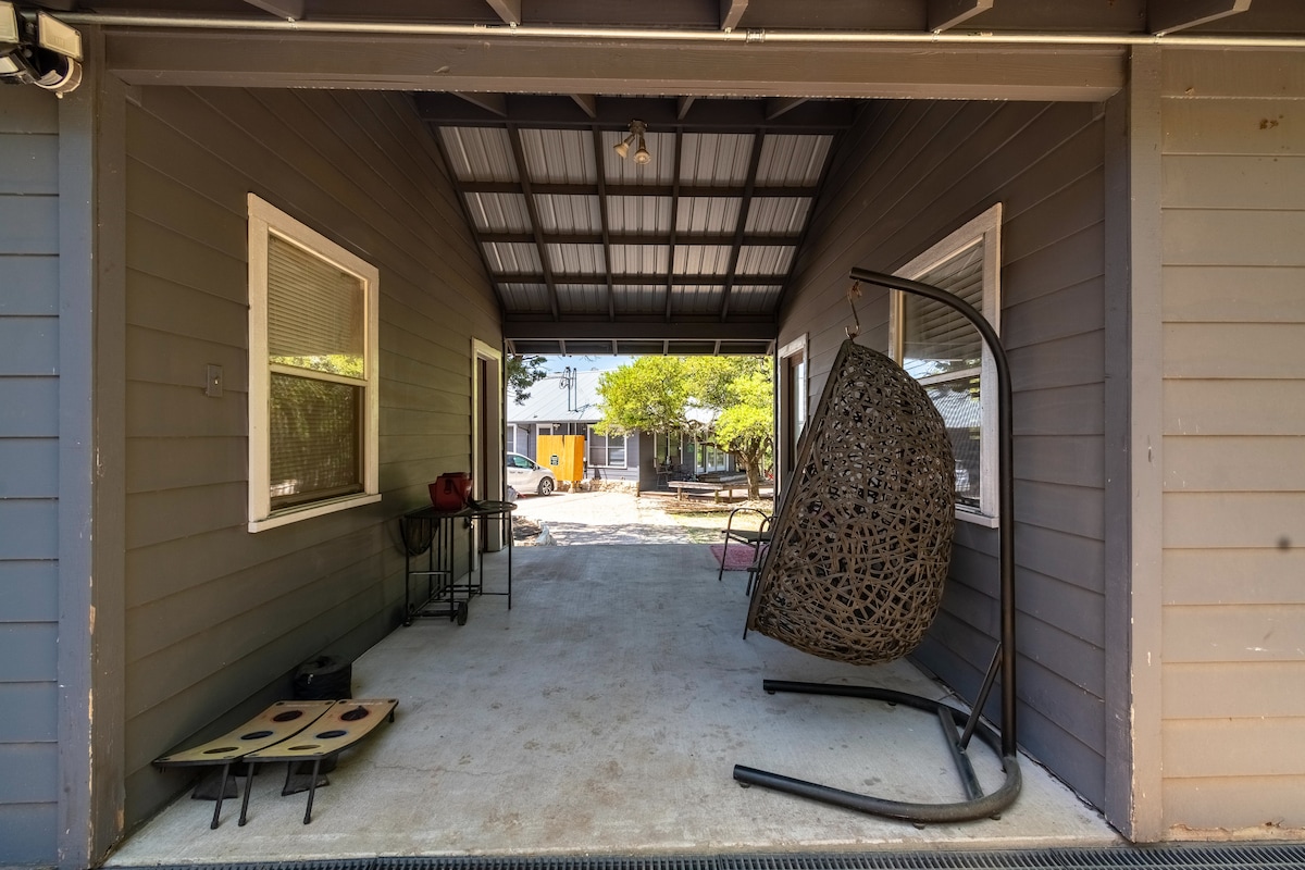 Covered porch at The Nook at Eco Valley featuring a hanging egg chair, shaded seating area, and view toward the shared grounds and cabins in a relaxed outdoor setting
