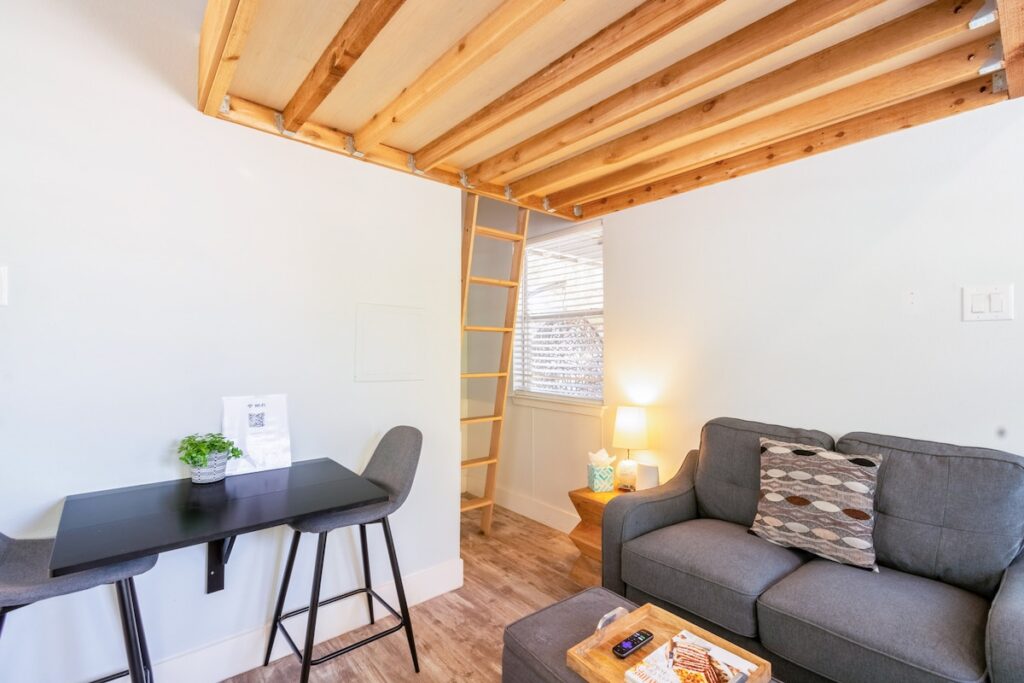 Bright interior of the Nook featuring a cozy sitting area with a gray sofa, small dining table, lofted wood ceiling with ladder access, and warm natural light creating a modern cabin feel