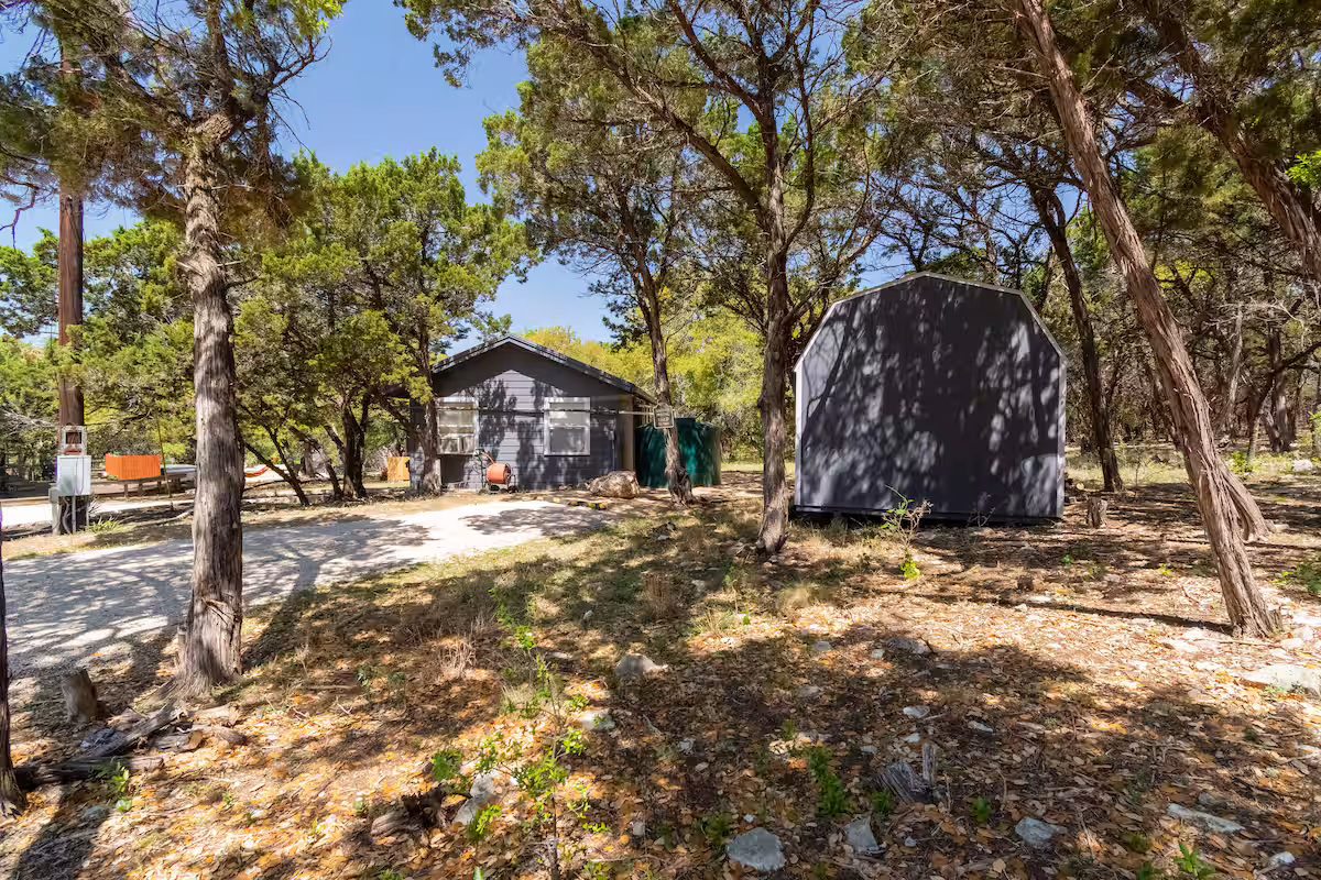 Small gray cabin and storage shed surrounded by cedar trees on a wooded eco property with a gravel path and natural landscape known as the Nook