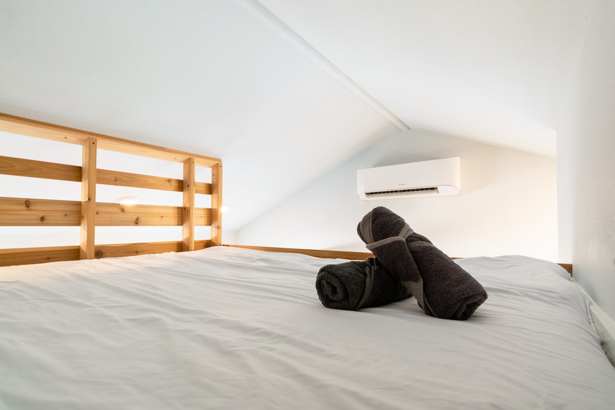 Loft sleeping area inside The Nook at Eco Valley with a comfortable bed, white linens, folded towels, wooden railing, and wall-mounted air conditioning for a cozy stay