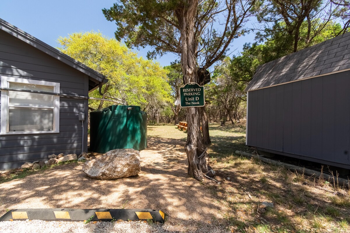 Reserved parking area for The Nook at Eco Valley with a Unit D sign mounted on a tree, shown between the cabin, storage shed, and rainwater tank in a wooded setting
