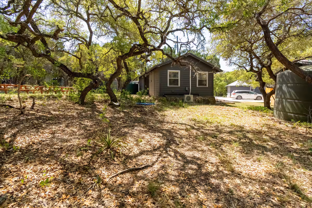 Wide exterior view of The Nook at Eco Valley nestled among oak trees, showing the cabin, natural ground cover, and surrounding wooded landscape with picnic area and rainwater tank