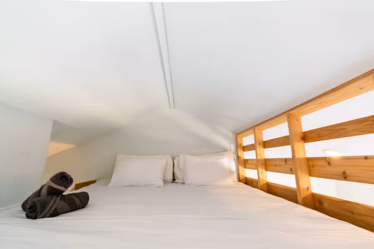 Loft bed inside The Nook at Eco Valley with white linens, wooden safety railing, folded towels, and soft natural light creating a cozy sleeping
