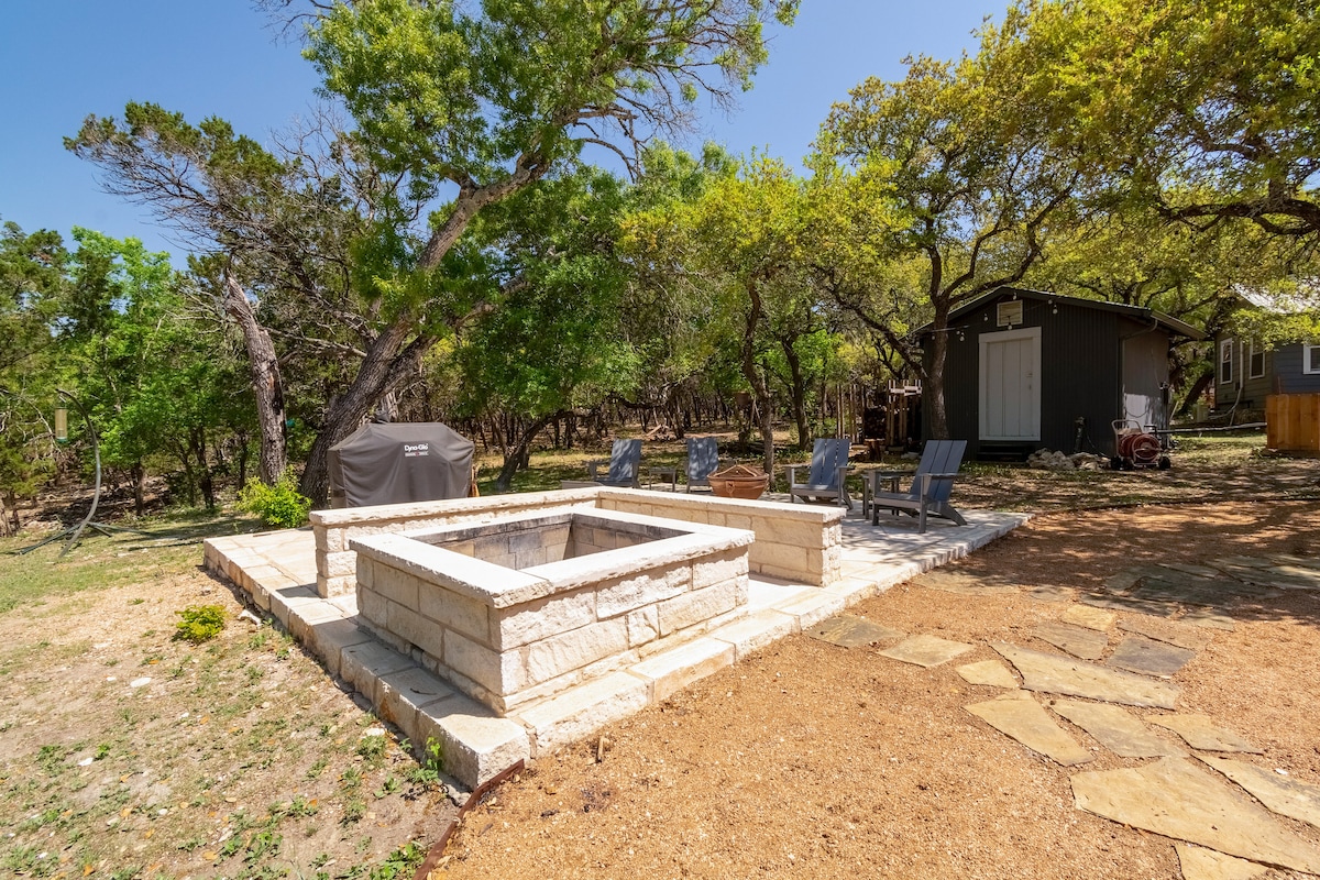 Outdoor fire pit area at The Nook at Eco Valley featuring a square stone fire pit, Adirondack chairs, grill, and shaded oak trees beside the cabin in a tranquil woodland setting