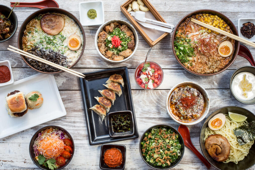 Overhead view of assorted Japanese dishes from Ramen Tatsuya, including ramen bowls with soft-boiled eggs, gyoza dumplings, rice bowls, sauces, and side dishes arranged on a wooden table.