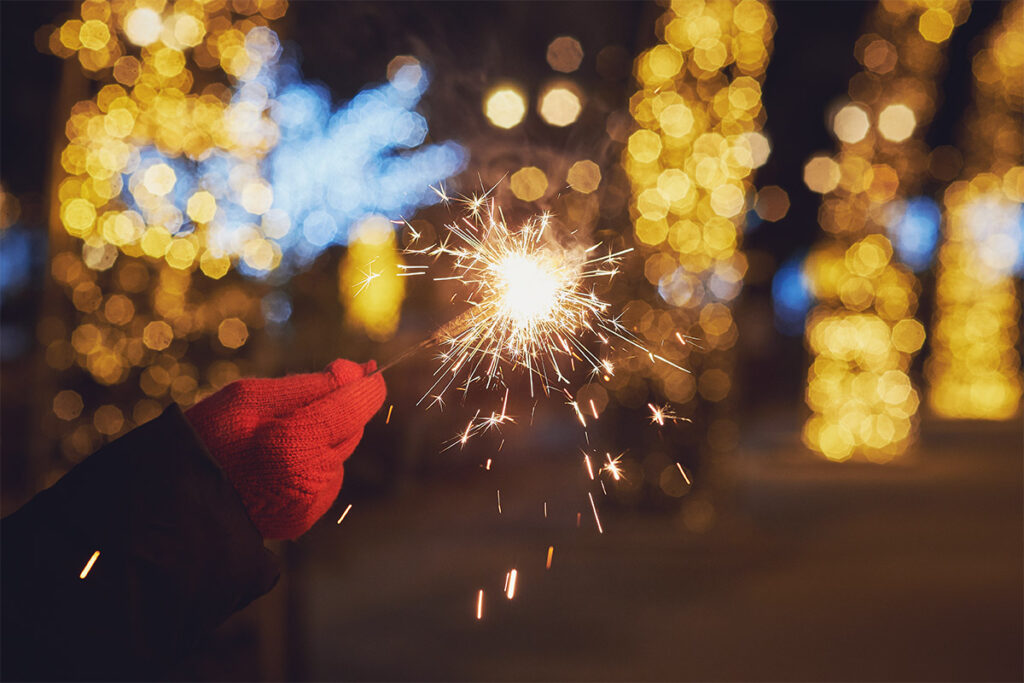 Hand holding a lit sparkler against a backdrop of festive holiday lights.