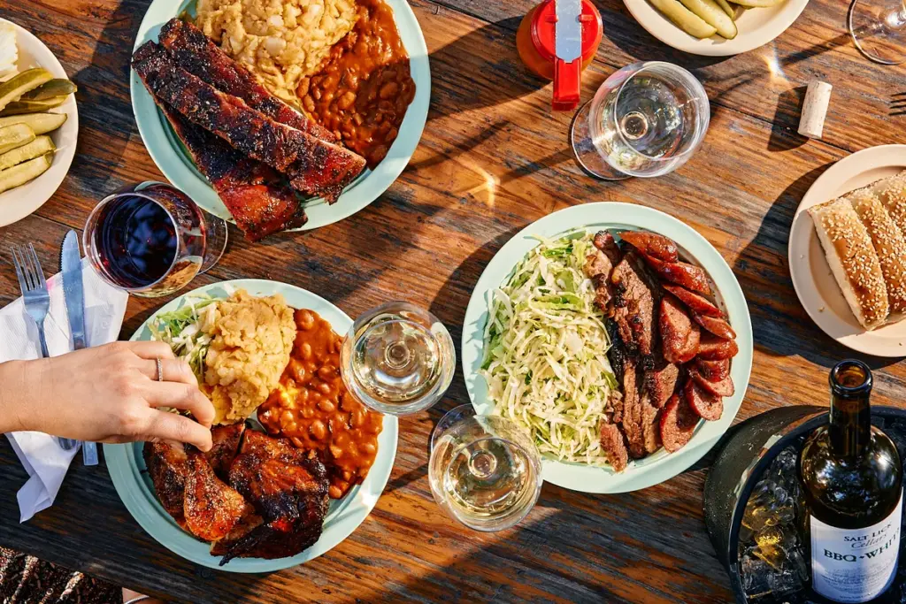 Plates of The Salt Lick BBQ featuring ribs, sausage, brisket, coleslaw, baked beans, and potato salad on a rustic wooden table with wine glasses and bottles.