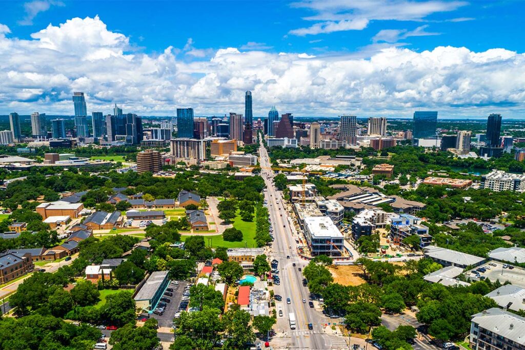 Aerial view of South Congress (SoCo) in Austin, Texas, with a long tree-lined roadway leading toward the downtown skyline under a bright blue sky with scattered clouds.